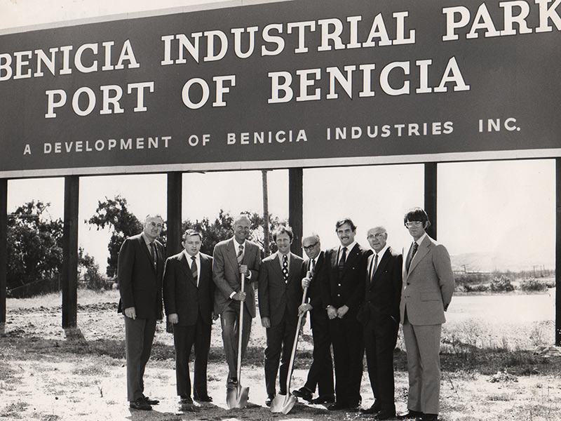 A sepia-toned photograph of eight men in suits, standing in front of a large billboard that says 'Benicia Industrial Park Port of Benicia'