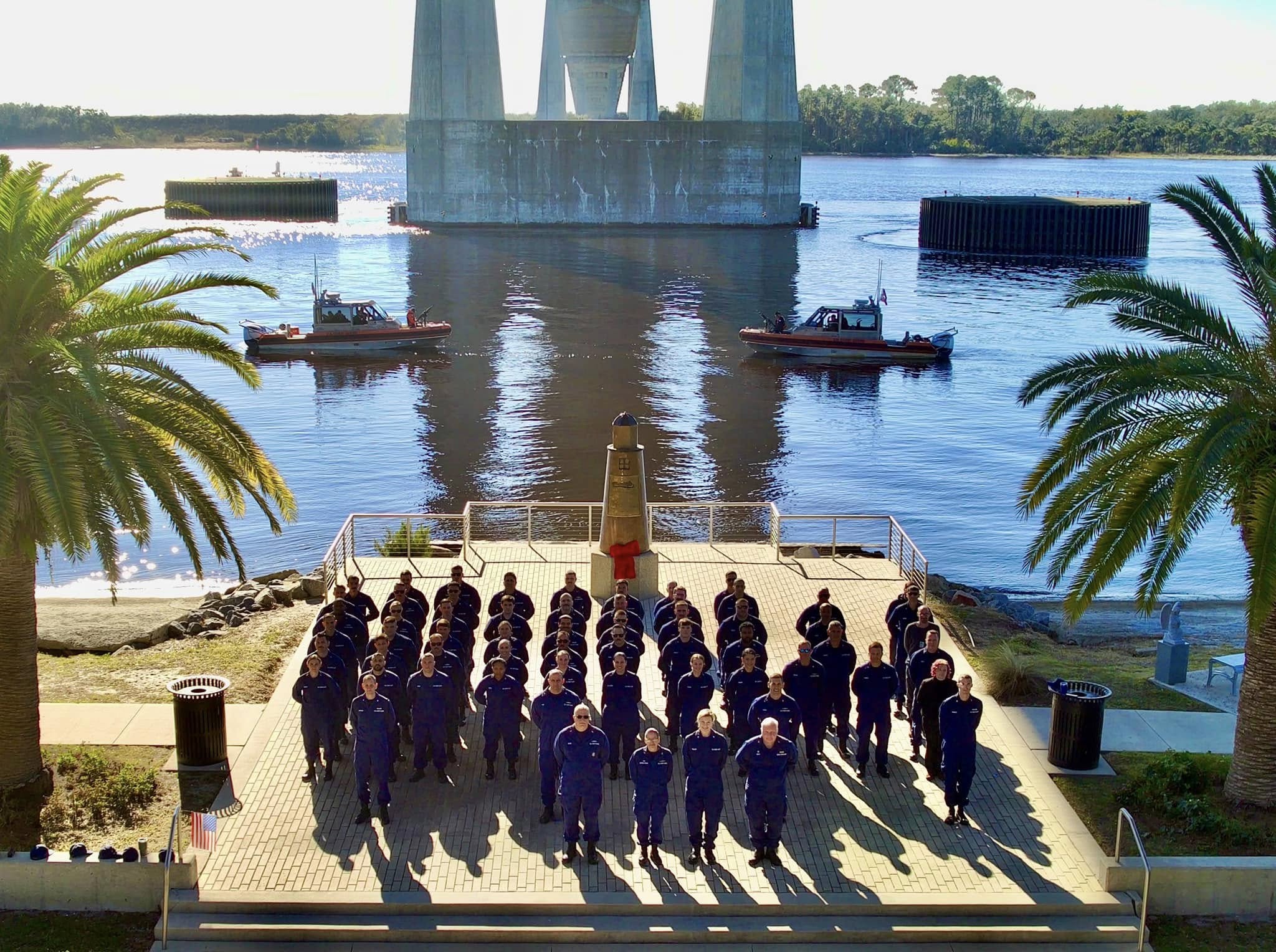 Group photo of Coast Guard personnel in dark blue uniforms standing in formation on a waterfront plaza
